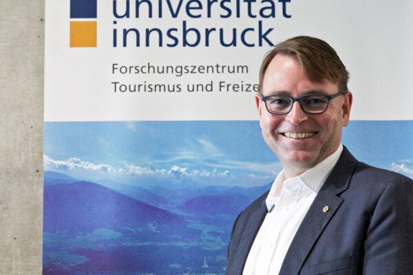 Alexander Plaikner Der Mann in Anzug und Brille präsentiert sich vor einem Hintergrund mit dem Logo der Universität Innsbruck und Berglandschaft.