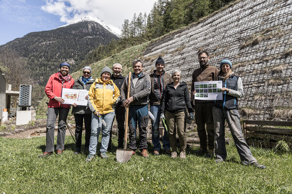 Kulturbotschafter Längenfeld Eine Gruppe von neun Personen steht auf einer Wiese, umgeben von Bergen, und hält Pläne für ein Projekt in den Händen.